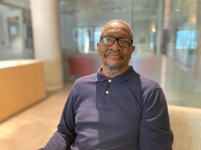 Photo of older Black man with a beard and mustache, wearing glasses sitting on a tan chair in foyer of apartment building